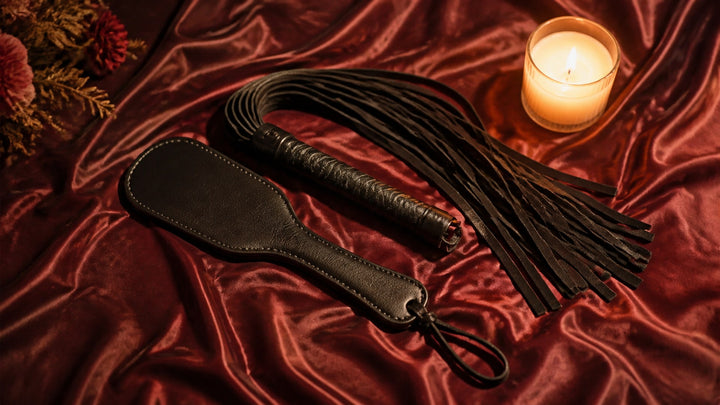 Black leather spanking paddle and fringe whip on maroon silk beside a lit candle