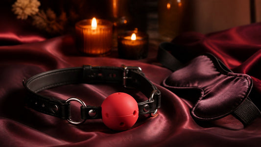 Red silicone ball gag with black strap on maroon silk beside a blindfold and candles
