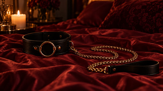 Black BDSM collar with a gold O-ring and chain leash on red silk sheets by candlelight