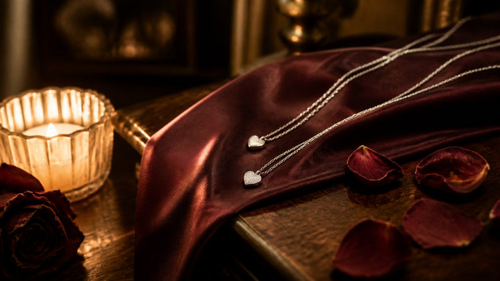 Two silver heart charm chains draped over maroon silk beside a warm candle, beginner nipple clamps mood setting
