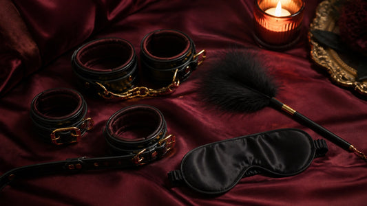 Couples bondage kit flat lay: faux-leather cuffs, satin blindfold and feather tickler on maroon silk