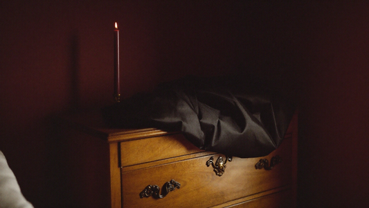 Moody intimate bedroom corner with soft black silk sash on a wooden dresser, single maroon candle, deep maroon wall in shadow