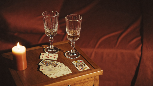 Warm intimate bedside scene with two crystal wine glasses, vintage card deck, and a single candle in deep maroon and amber tones