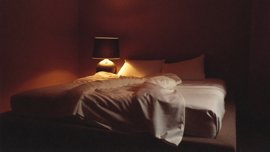 Dimly-lit intimate bedroom at dawn, soft maroon lamplight on rumpled white linen sheets