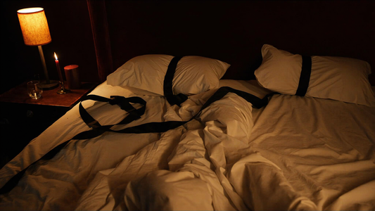 Cinematic boudoir close-up of a black silk restraint sash draped over rumpled white sheets with a single maroon candle and empty wine glass, deep amber lamplight
