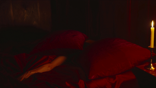 Cinematic intimate bedroom scene with two crimson velvet pillows entangled on rumpled wine-red silk sheets, single tall candle, deep maroon and amber moody atmosphere