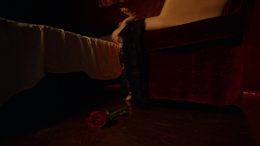 Cinematic intimate boudoir scene with a delicate black lace slip dress draped over a deep maroon velvet armchair beside a single fresh maroon rose, candlelit deep night atmosphere