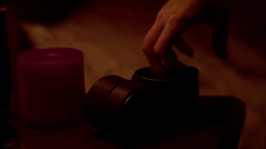 Cinematic intimate close-up of soft black leather wrist cuffs resting open beside a single low-lit maroon candle on dark walnut nightstand, silhouette of a man's fingertips reaching from off-frame, deep amber and maroon mood