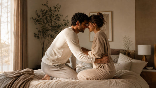 Couple in soft loungewear kneeling close on a bed in warm morning light