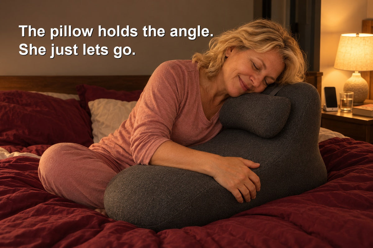 A woman in her fifties sitting on a dark red duvet and hugging the gray WildNightX Pillow, with the words: The pillow holds the angle. She just lets go.