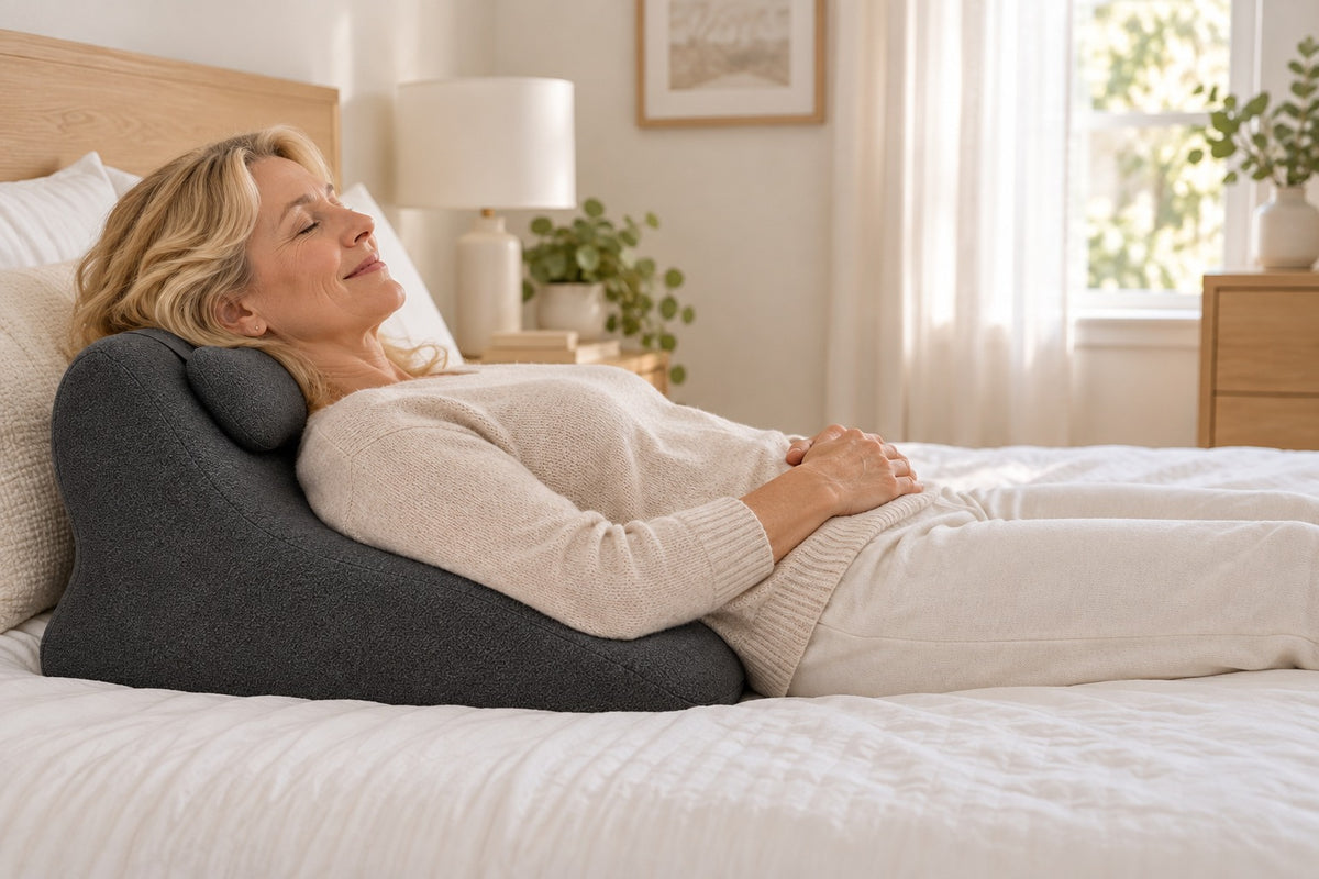 Woman relaxing in bed with her eyes closed, leaning back on the WildNightX Pillow propped against the headboard